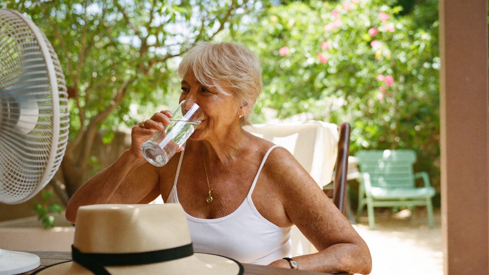 Senior woman staying cool at home with a glass of water and a fan on a hot summer day